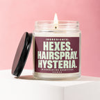 Glass candle with burning wick and lid, featuring label text: "Ingredients: Hexes. Hairspray. Hysteria. In Escalating Quantities. Vowed."