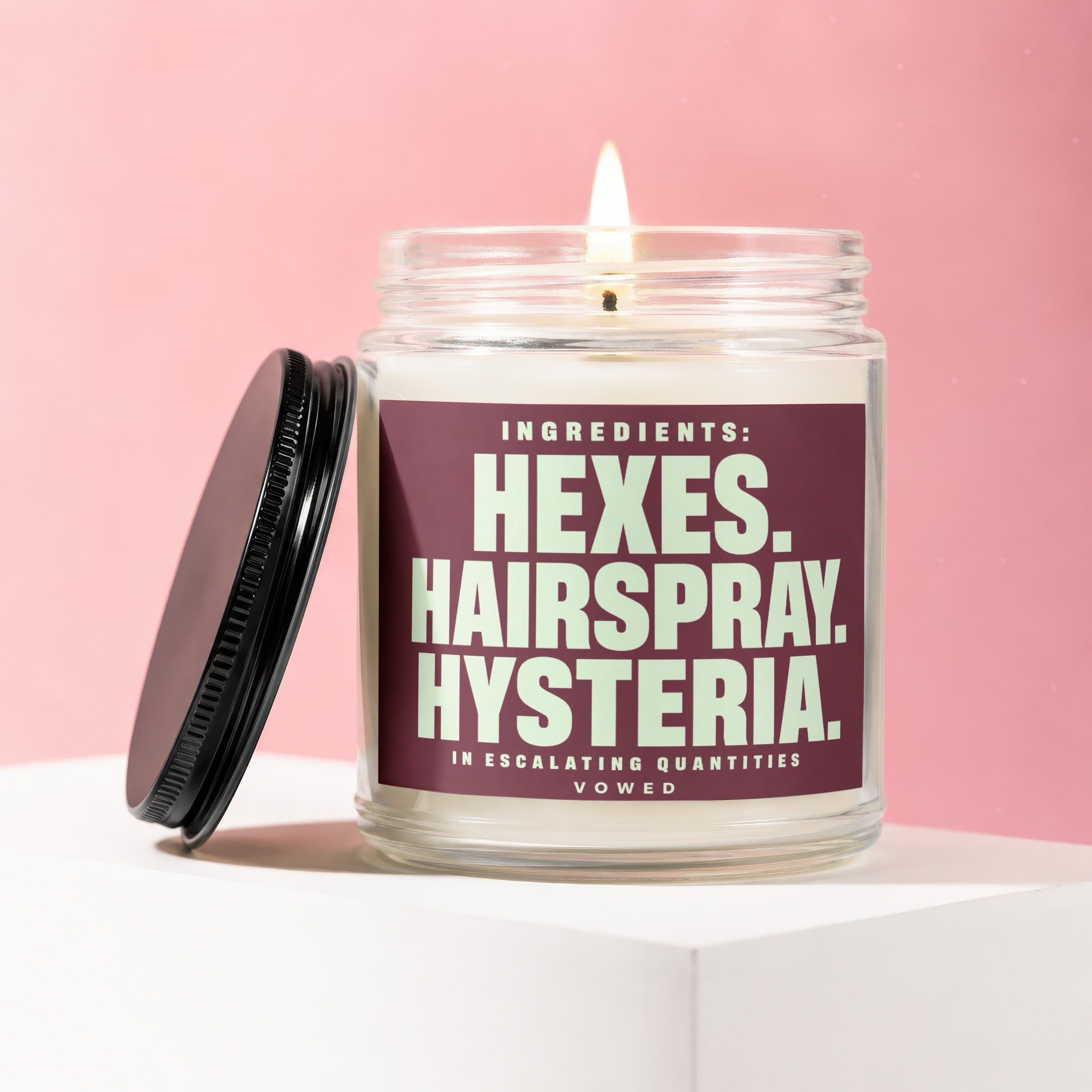 Glass candle with burning wick and lid, featuring label text: "Ingredients: Hexes. Hairspray. Hysteria. In Escalating Quantities. Vowed."