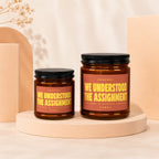 Two amber candles with black lids and pink labels featuring yellow text.