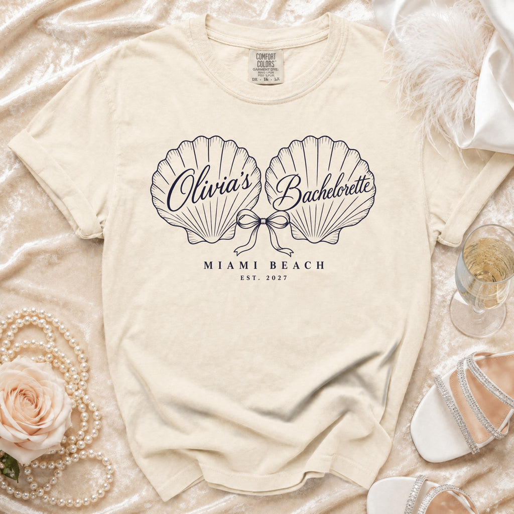 A cream-colored t-shirt with two seashell illustrations, a bow, and text that reads Olivia's Bachelorette Miami Beach Est. 2027.