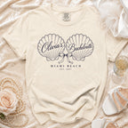 A cream-colored t-shirt with two seashell illustrations, a bow, and text that reads Olivia's Bachelorette Miami Beach Est. 2027.