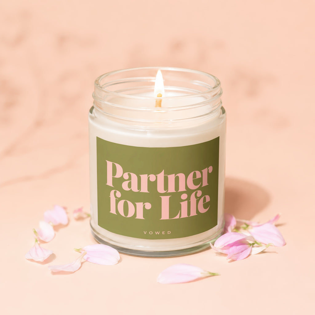 Candle with green label reading "Partner for Life VOWED" surrounded by pink petals.