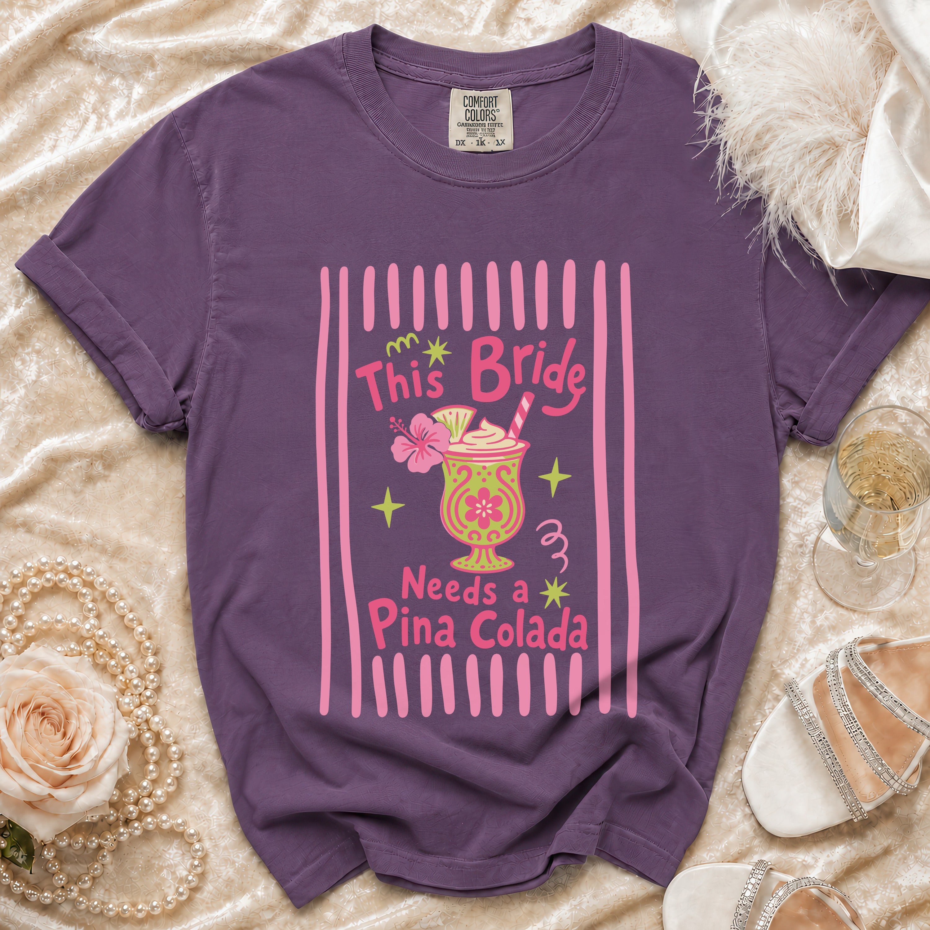 Purple t-shirt with pink text "This Bride Needs a Pina Colada" and tropical drink graphic.