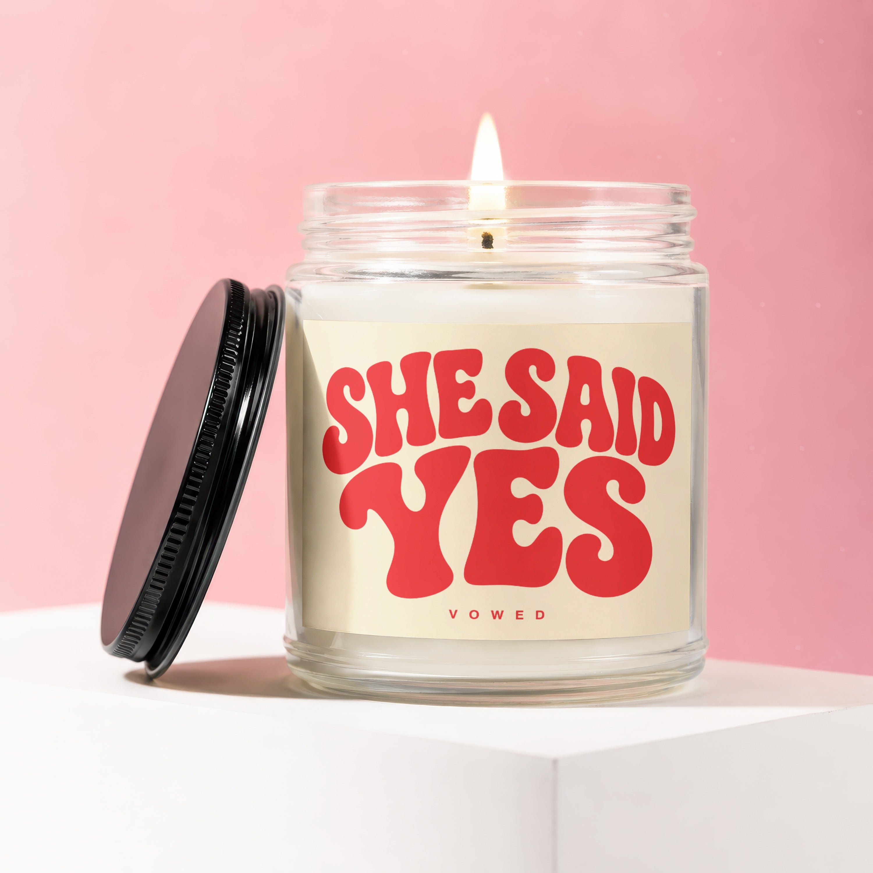 Glass jar candle with a lit wick and red text that reads "SHE SAID YES VOWED."