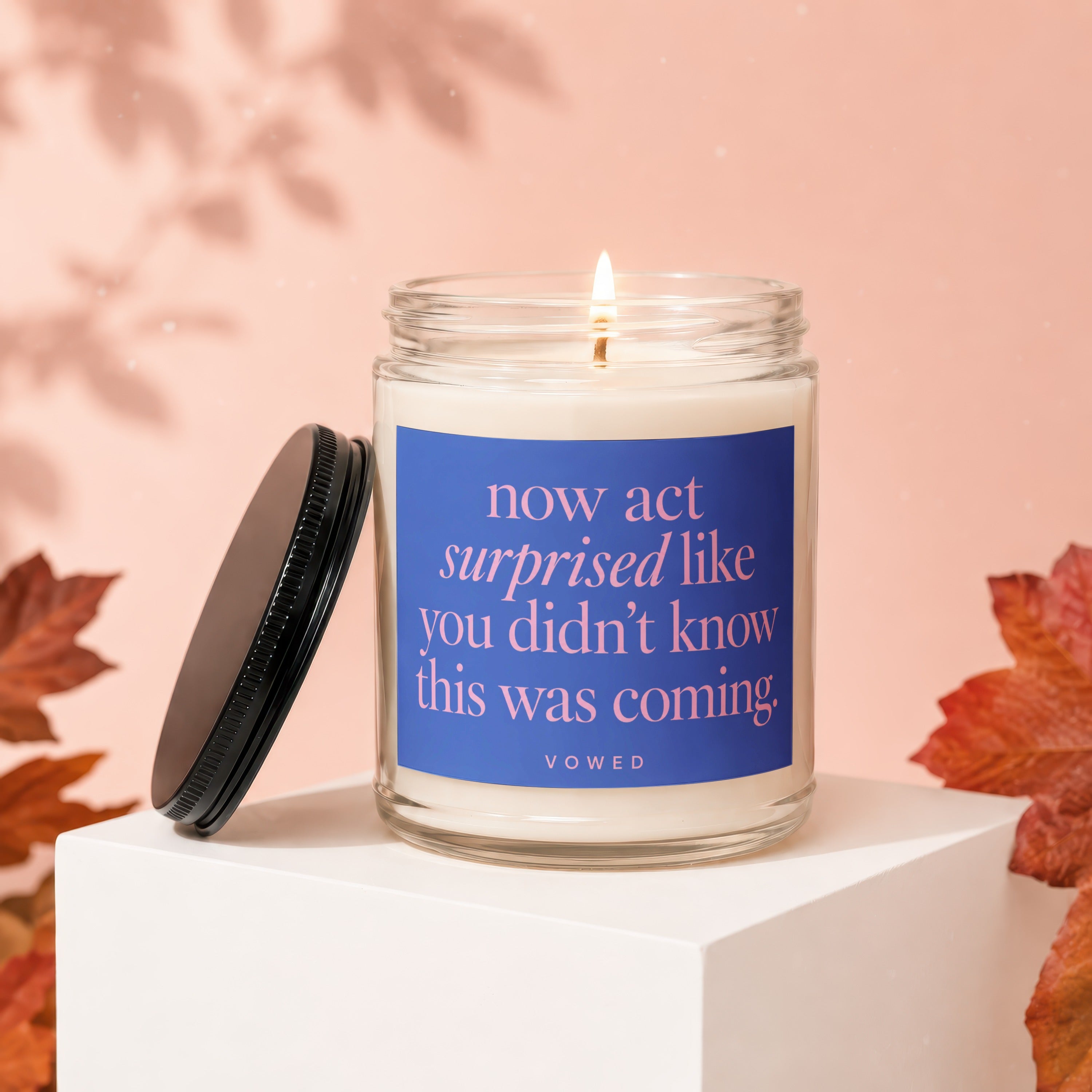 Glass candle with lit wick, blue label reads "now act surprised like you didn't know this was coming. VOWED".