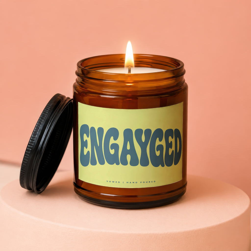 Amber jar candle with lid off, displaying "ENGAYGED" text and "Vowed | Hand Poured" label.