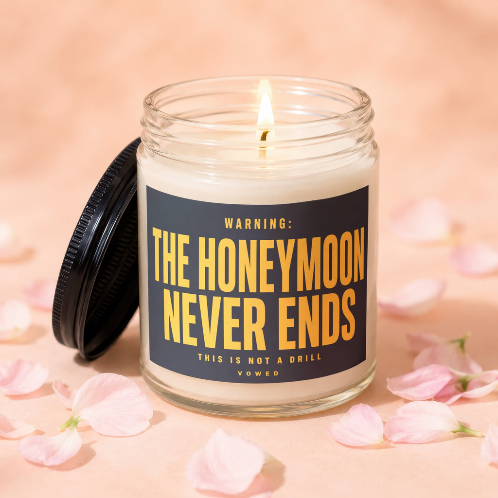 Glass candle with lit wick, lid off. Label reads "WARNING: THE HONEYMOON NEVER ENDS".