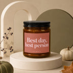 Candle with "Best day, best person" text on a peach label, on a display stand.
