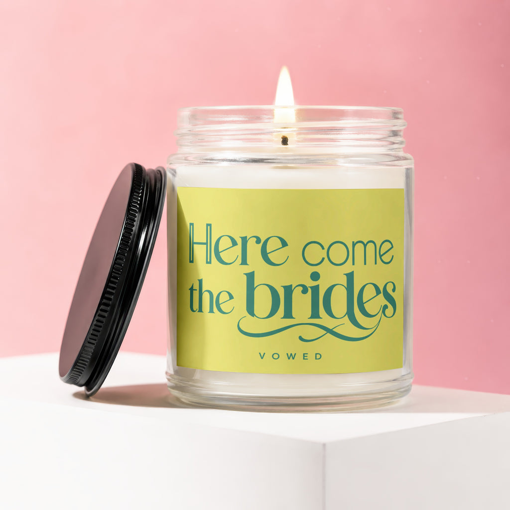 Lit candle in glass jar with black lid, reading "Here come the brides VOWED".
