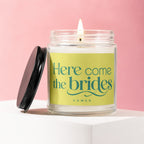 Lit candle in glass jar with black lid, reading "Here come the brides VOWED".