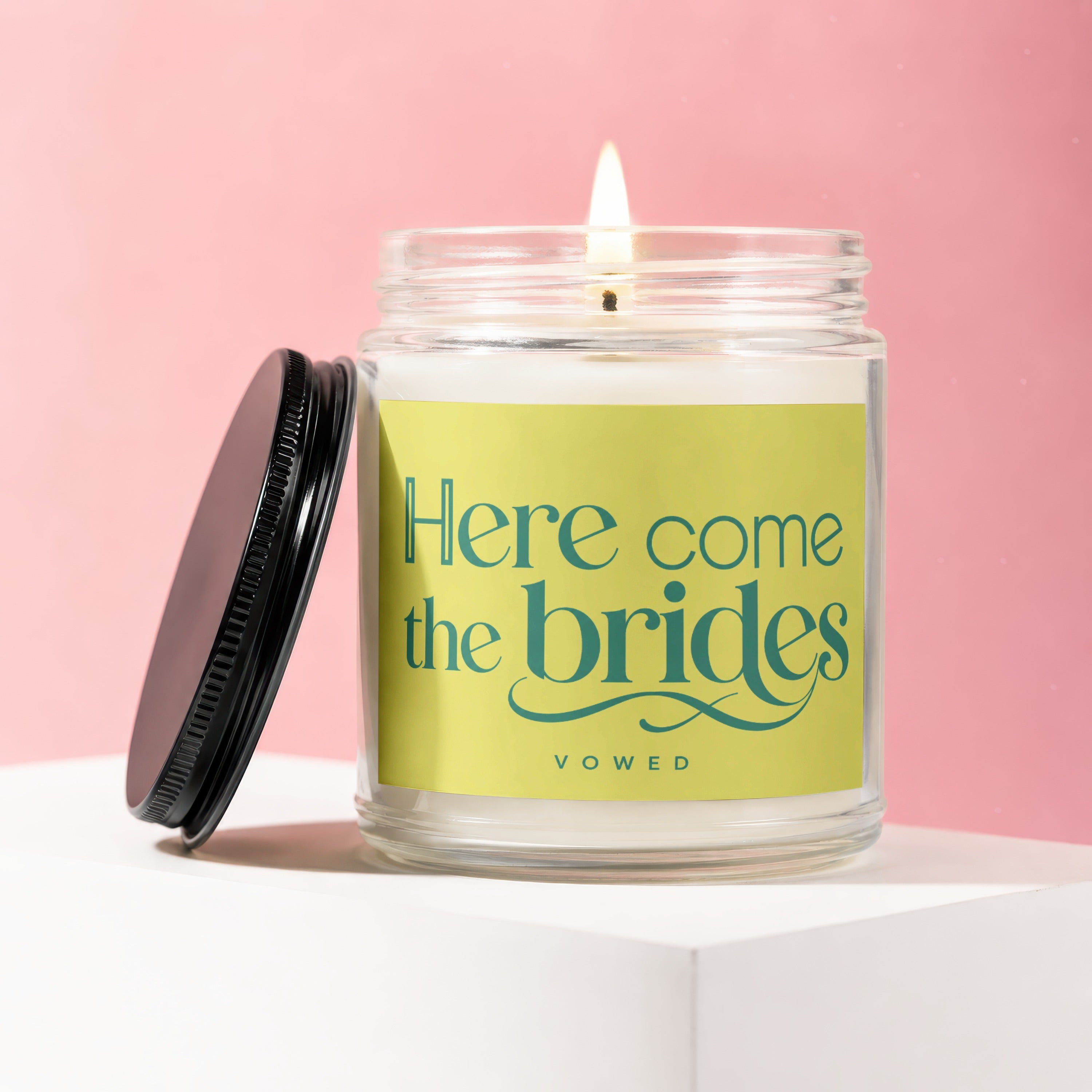Lit candle in glass jar with black lid, reading "Here come the brides VOWED".