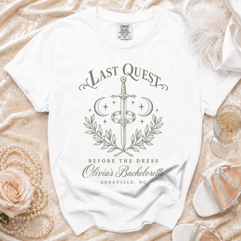 White t-shirt featuring green fantasy-themed text that reads Last Quest Before the Dress and Olivia's Bachelorette Asheville, NC.