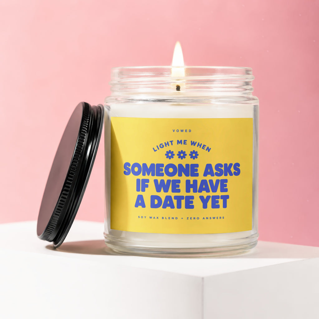 Lit candle with yellow label reading "Light me when someone asks if we have a date yet."