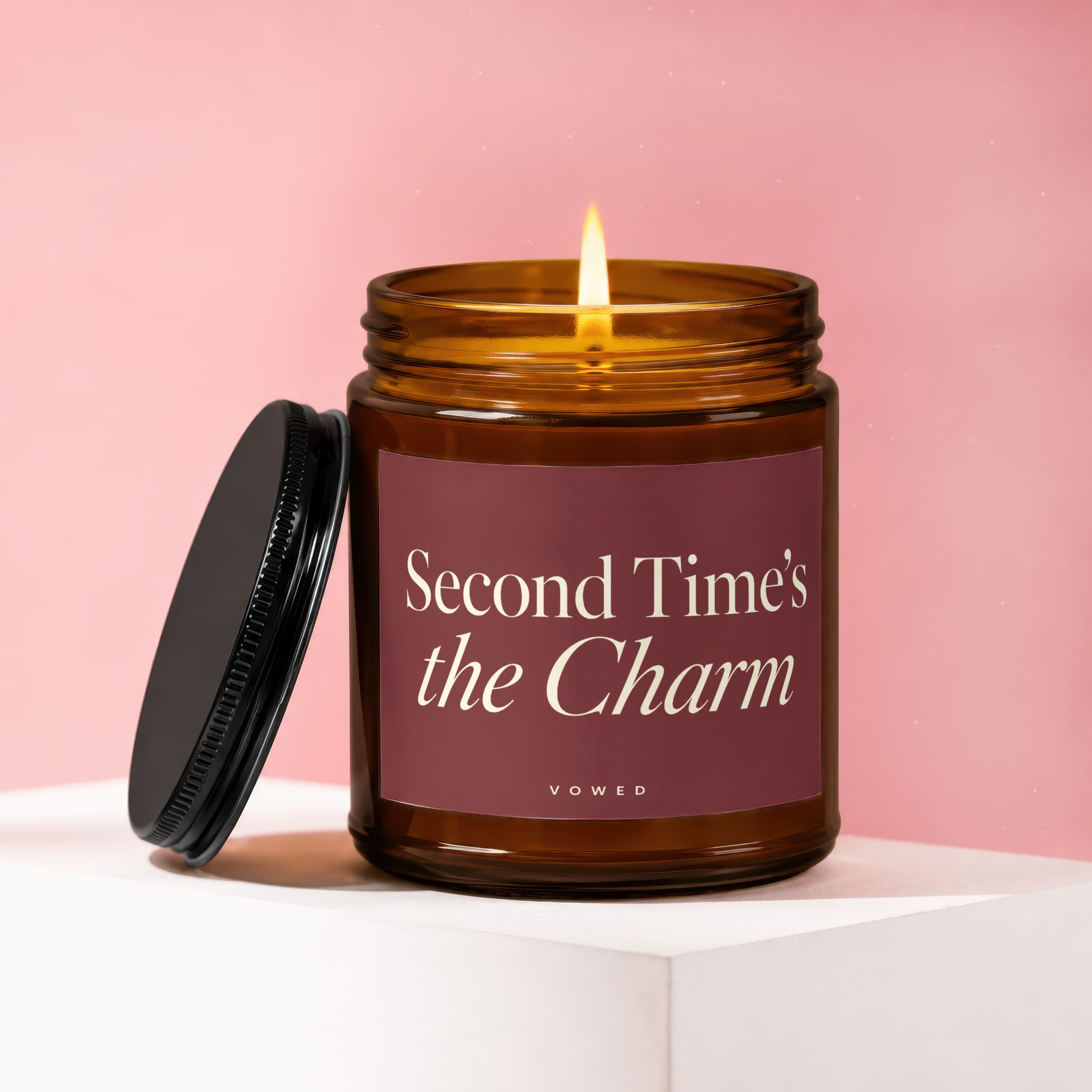 Amber glass candle with lit flame and lid removed, on display. Label reads "Second Time's the Charm."
