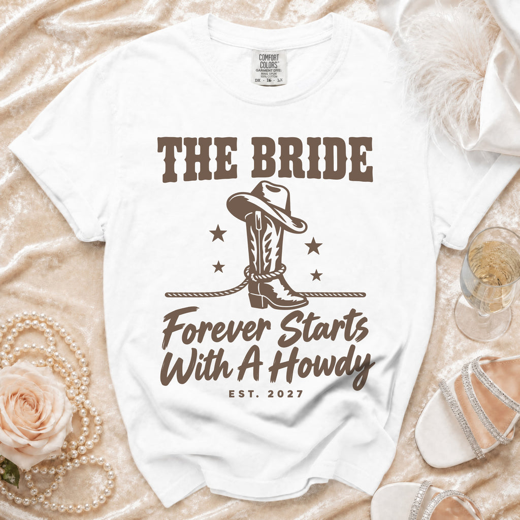 A white t-shirt featuring brown text reading "THE BRIDE", a western boot with a cowboy hat and stars, and "Forever Starts With A Howdy Est. 2027".