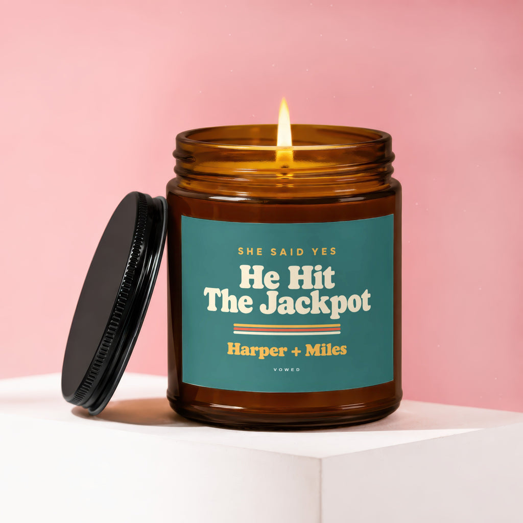 Amber jar candle with burning wick and black lid, "He Hit The Jackpot" text.