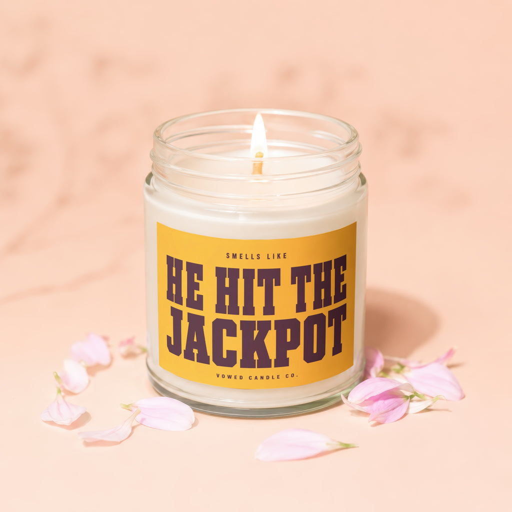 Glass jar candle with lit wick, reads "Smells Like He Hit The Jackpot."