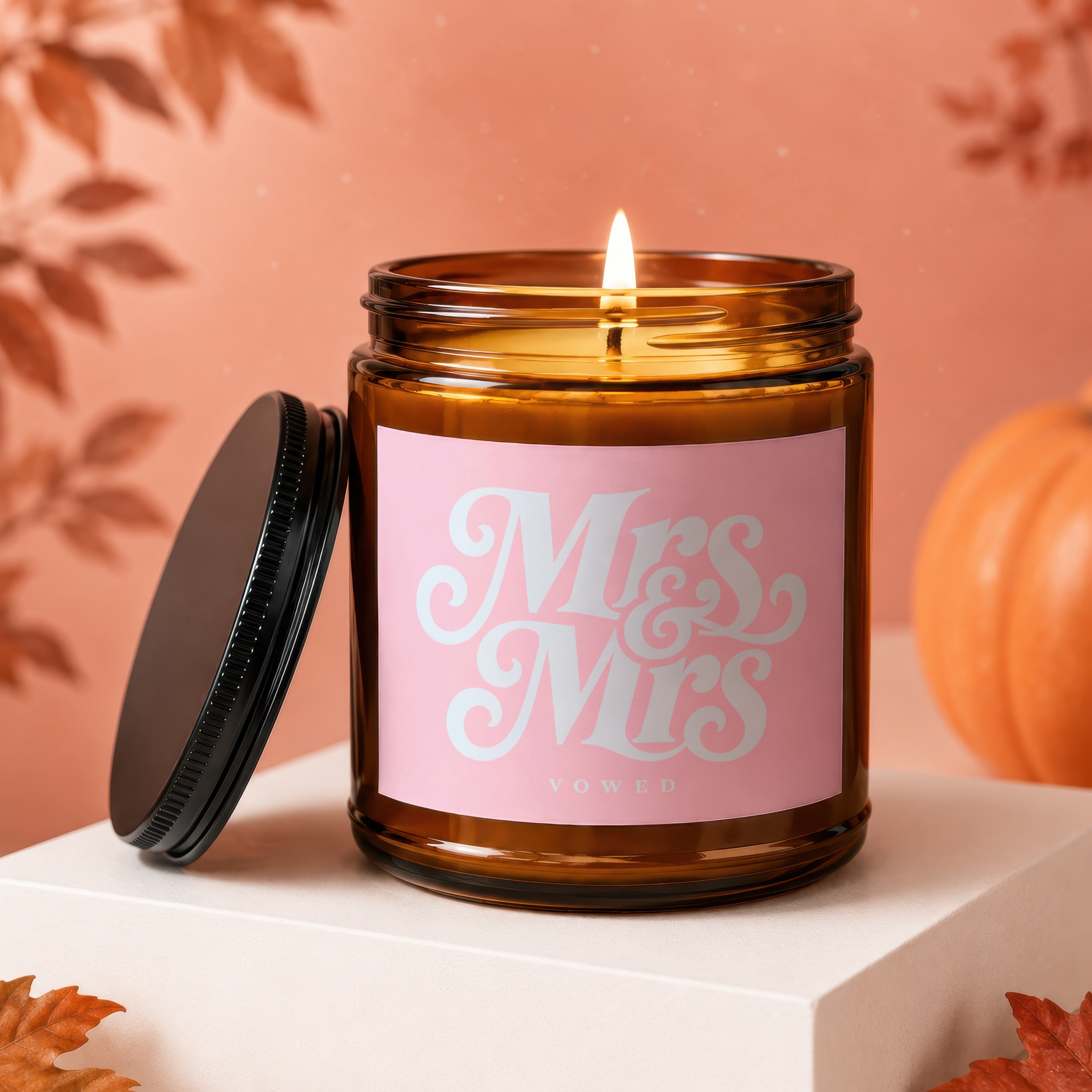 Amber jar candle with pink label reading "Mrs & Mrs VOWED," lit with lid beside it.