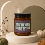 Brown candle in amber jar with "You're one lucky b*tch" label and black lid.