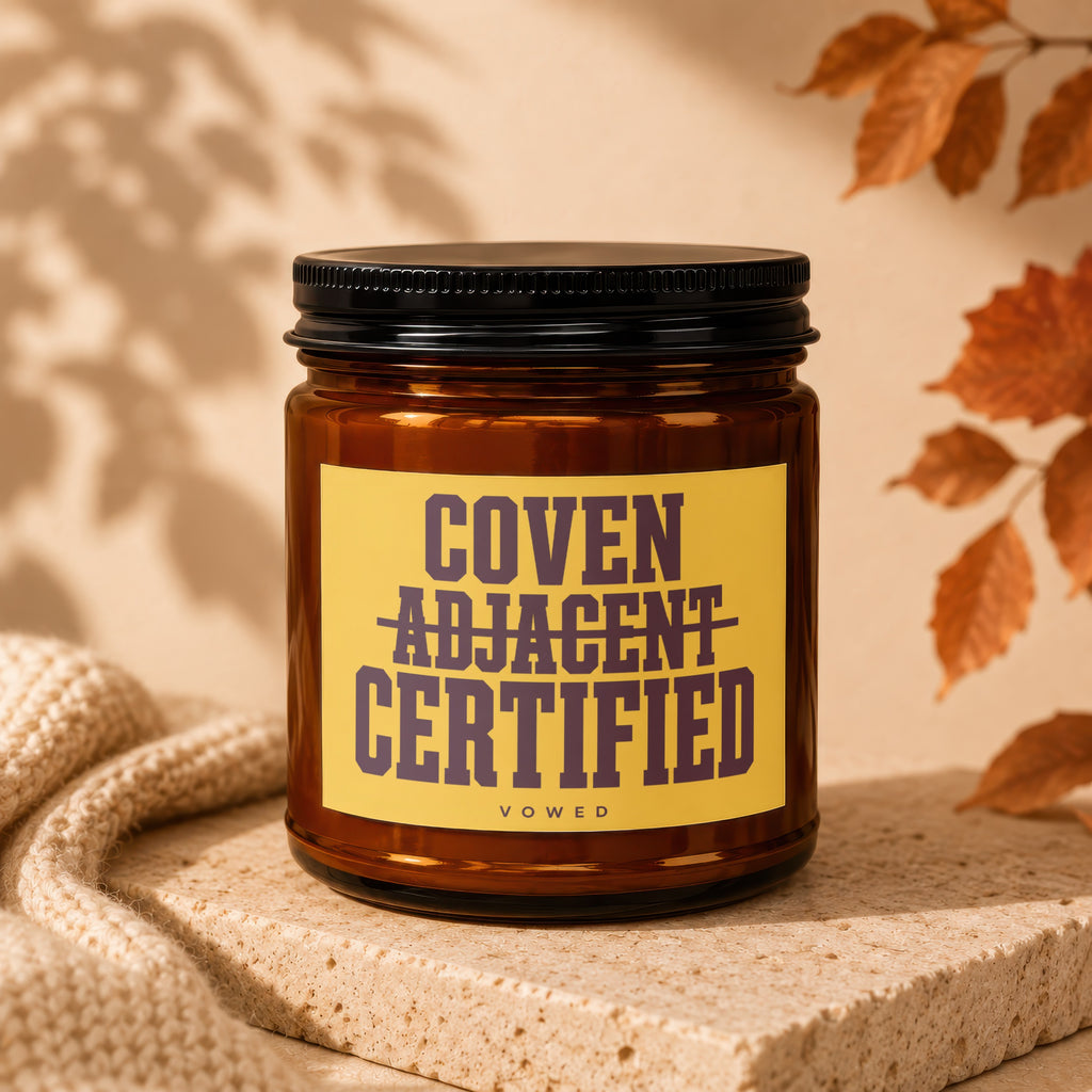 Amber candle with black lid and yellow label reading "COVEN ADJACENT CERTIFIED VOWED".