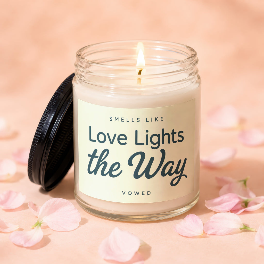 Lit candle in glass jar with black lid, text reads "Smells Like Love Lights the Way Vowed".