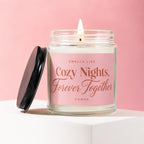 Candle with a lit wick and a pink label reading "Smells Like Cozy Nights, Forever Together".