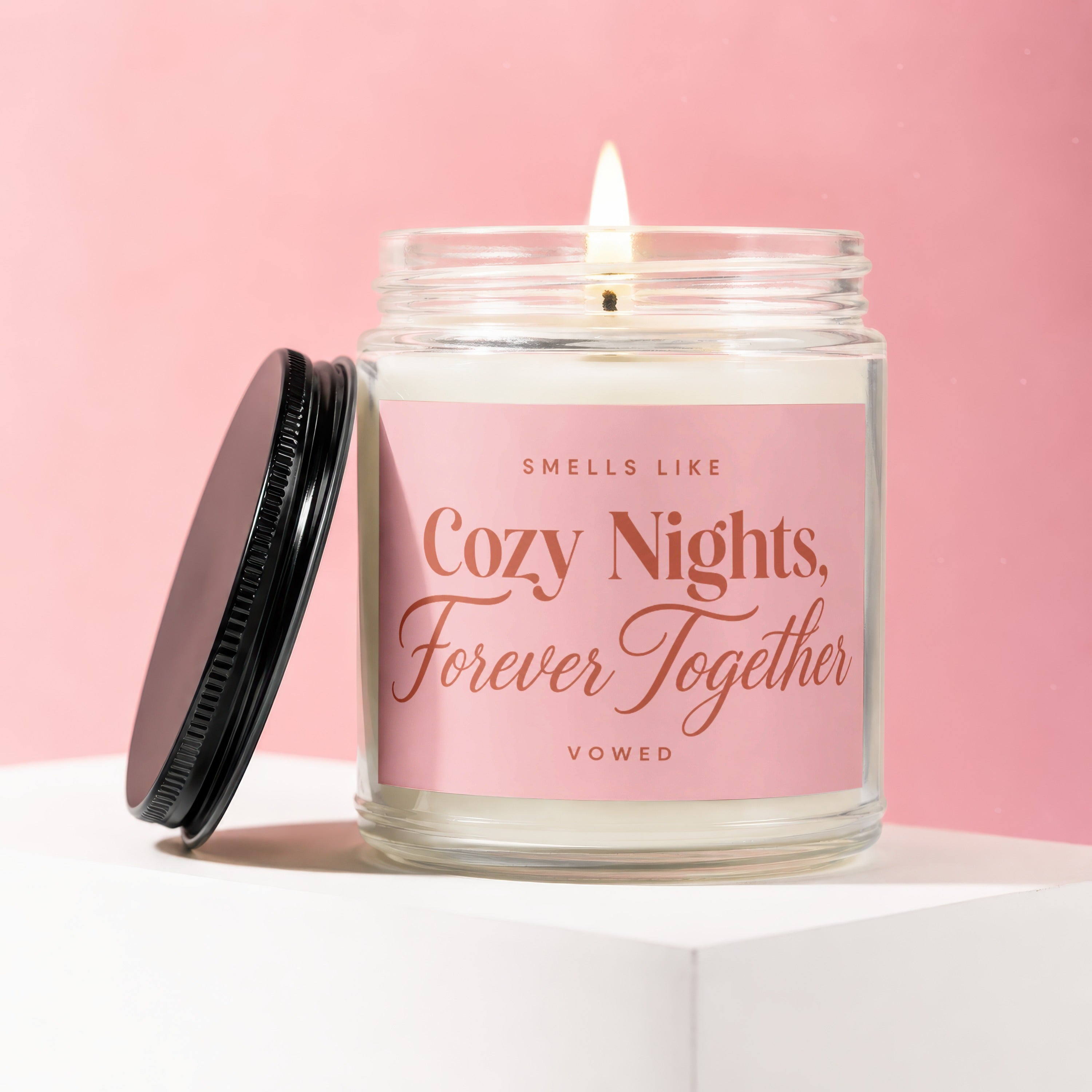 Candle with a lit wick and a pink label reading "Smells Like Cozy Nights, Forever Together".