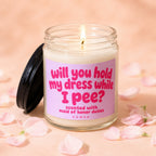 Glass candle with pink label that reads "Will you hold my dress while I pee?"