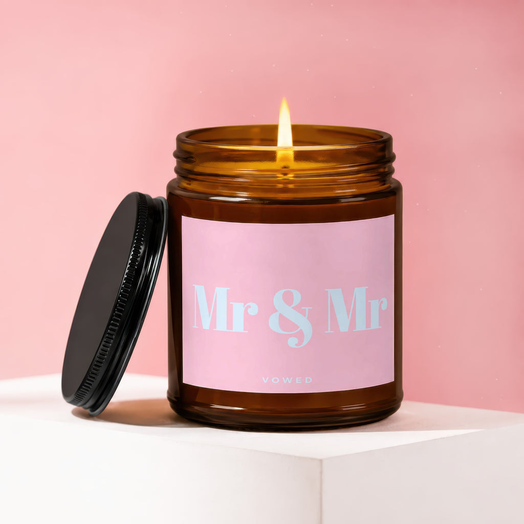 Amber jar candle with lit flame and "Mr & Mr VOWED" text on a pink background.