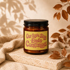 Amber candle with a yellow label reading "Our Love Story Ends Continues" on a stone riser.