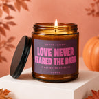 Amber jar candle with lit flame reads "Love Never Feared The Dark".