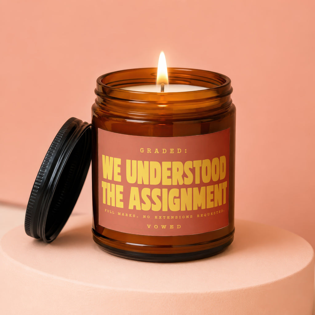 Amber jar candle with lit flame and label reading "We Understood The Assignment."