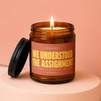 Amber jar candle with lit flame and label reading "We Understood The Assignment."
