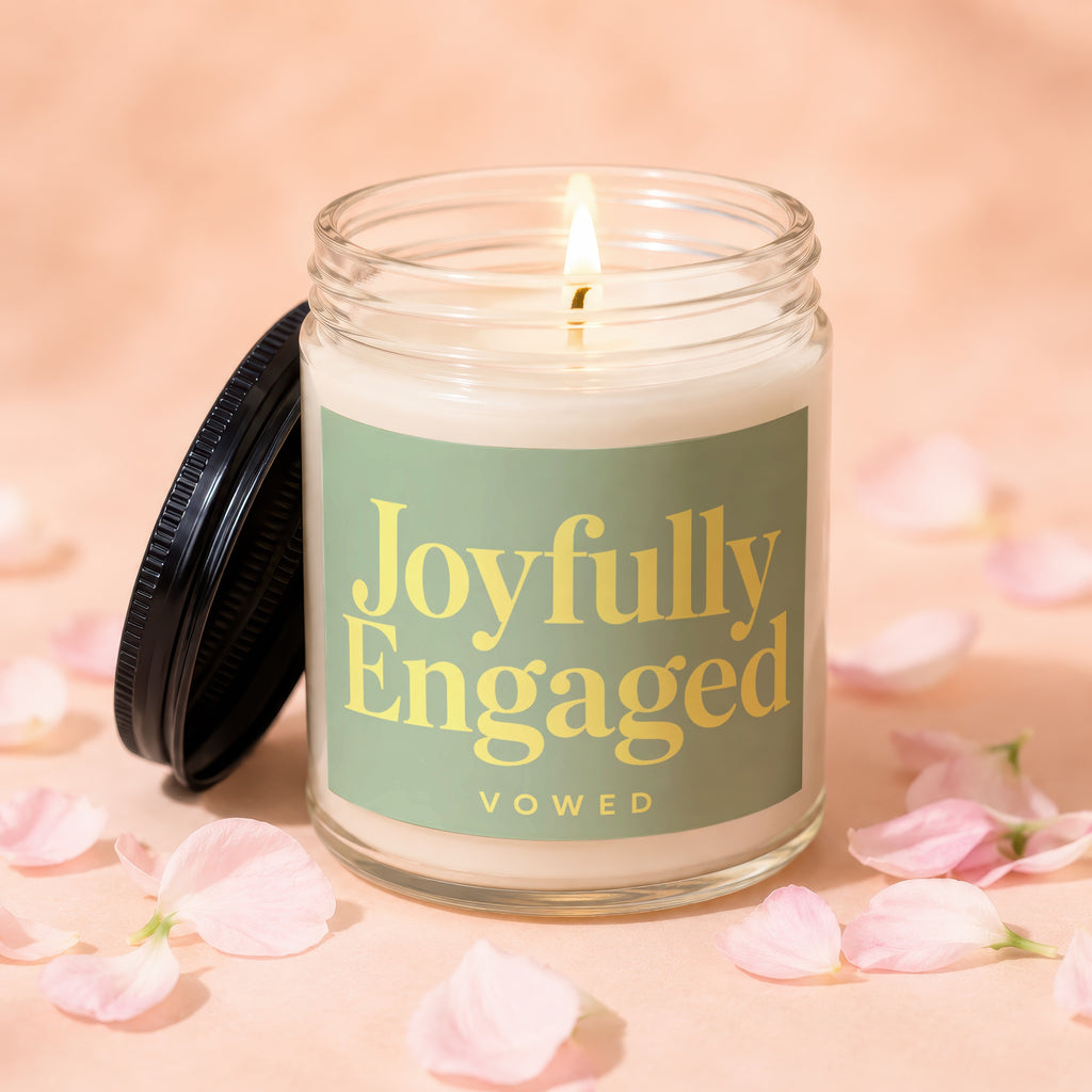 Glass candle with lit wick on a pink surface, surrounded by pink petals.