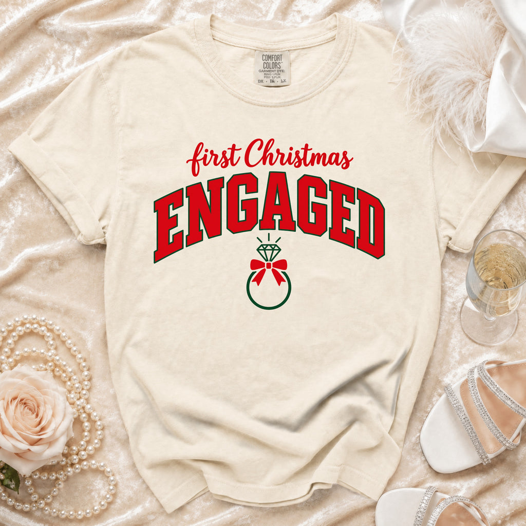 Cream-colored t-shirt featuring red and green text that reads first Christmas ENGAGED above a ring illustration.