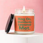 Glass candle with lit wick, orange label reads "Ring On. Standards Lowered Met. VOWED | HAND POURED."