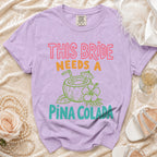 Purple t-shirt with "THIS BRIDE NEEDS A PINA COLADA" graphic and coconut drink.