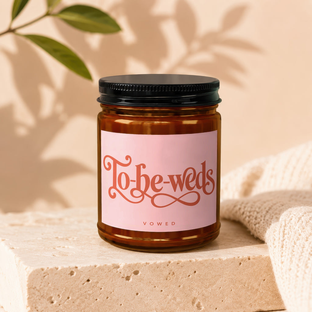 Candle with "To-be-weds" and "VOWED" text on pink label, amber glass jar with black lid.