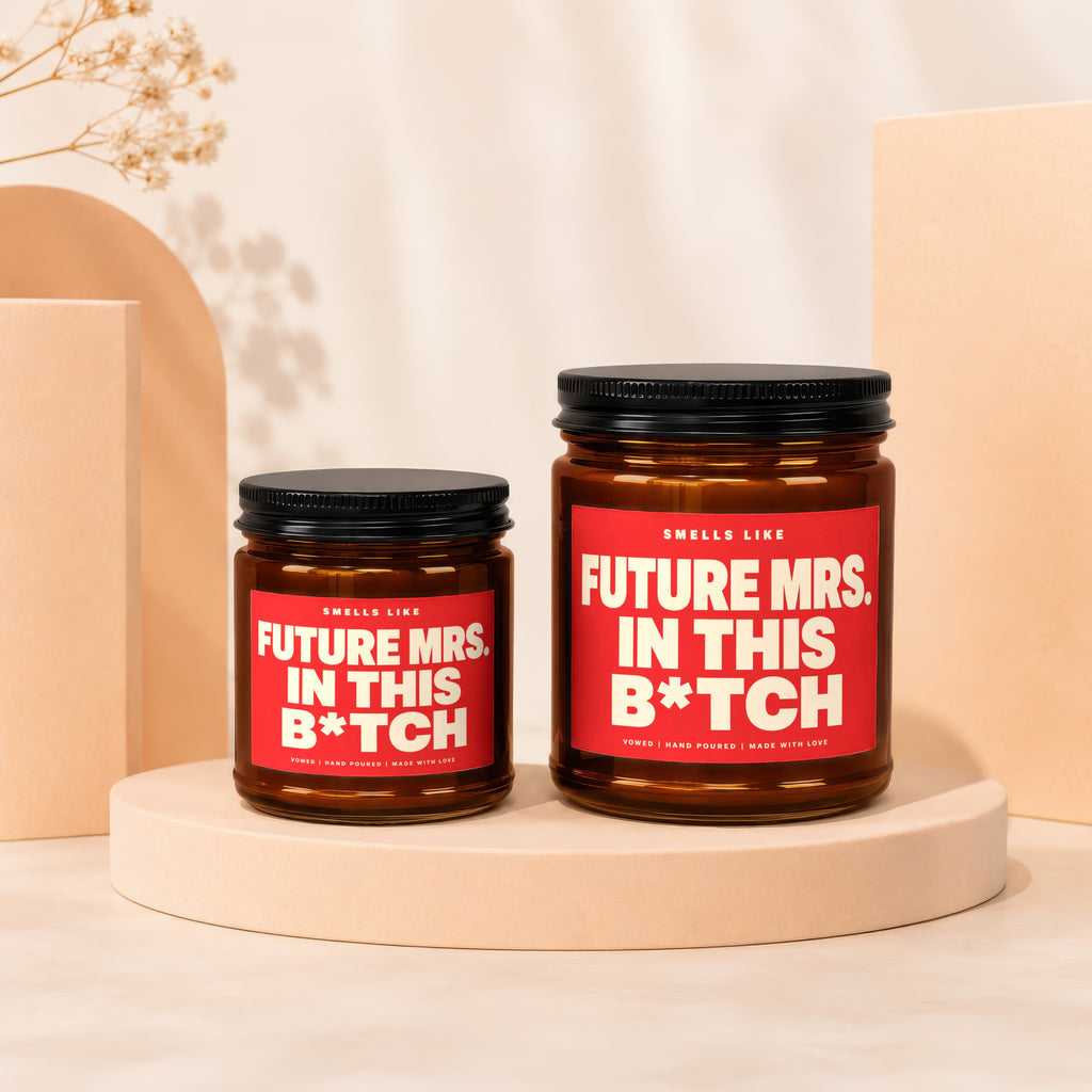Two amber candles with red labels reading "Smells Like Future Mrs. In This B*tch".