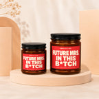 Two amber candles with red labels reading "Smells Like Future Mrs. In This B*tch".