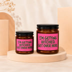 Two amber glass candles with black lids, featuring pink labels with green text.