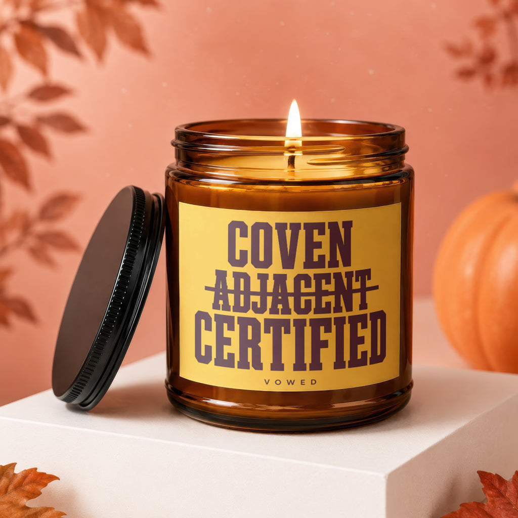 Amber jar candle with lit flame and yellow label reading "Coven Adjacent Certified".