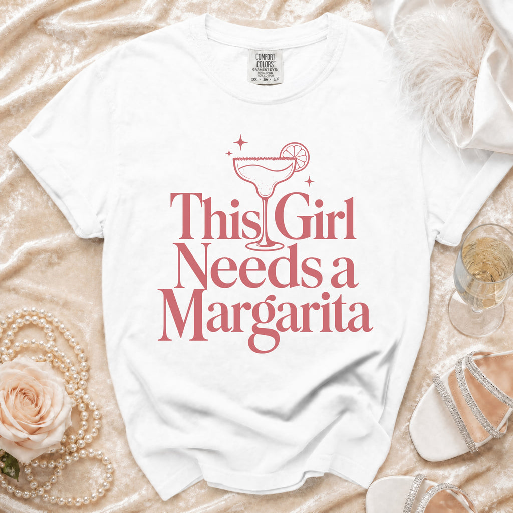 White t-shirt with pink text and margarita graphic that reads "This Girl Needs a Margarita".