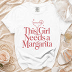 White t-shirt with pink text and margarita graphic that reads "This Girl Needs a Margarita".