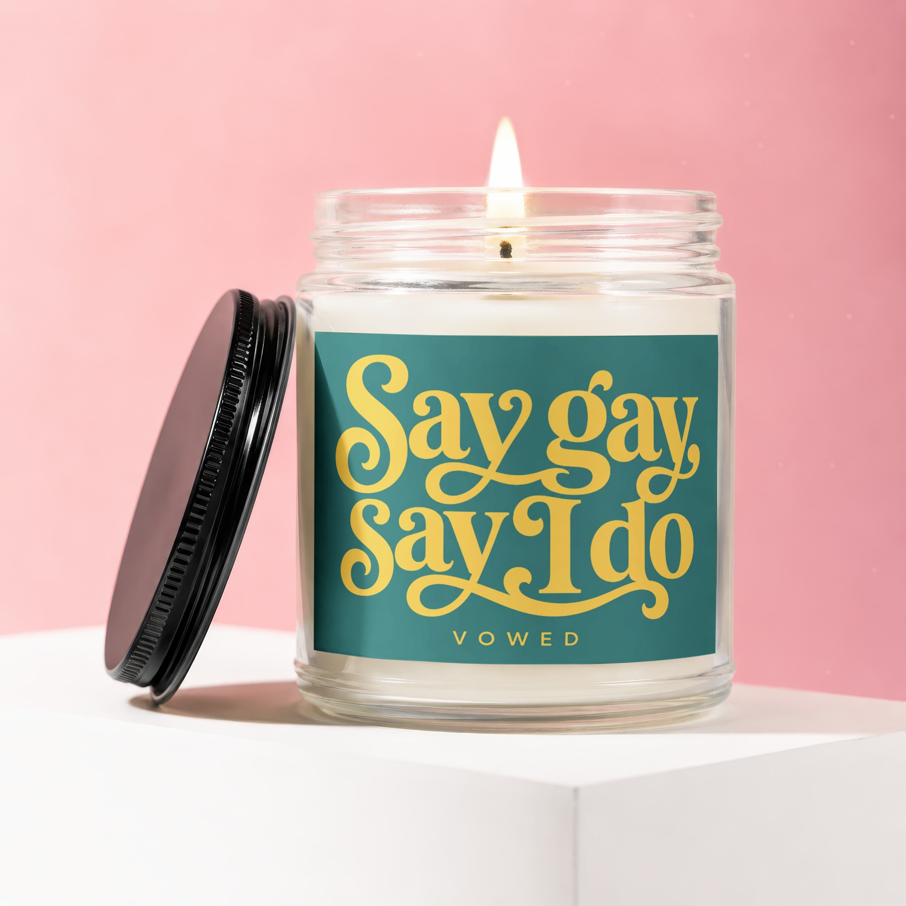 Jar candle with lit wick, featuring a teal label with yellow text "Say gay Say I do VOWED."