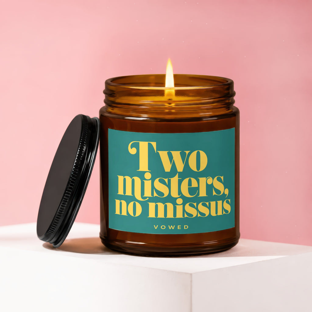 Amber jar candle with burning wick, lid off. Label reads "Two misters, no missus vowed".
