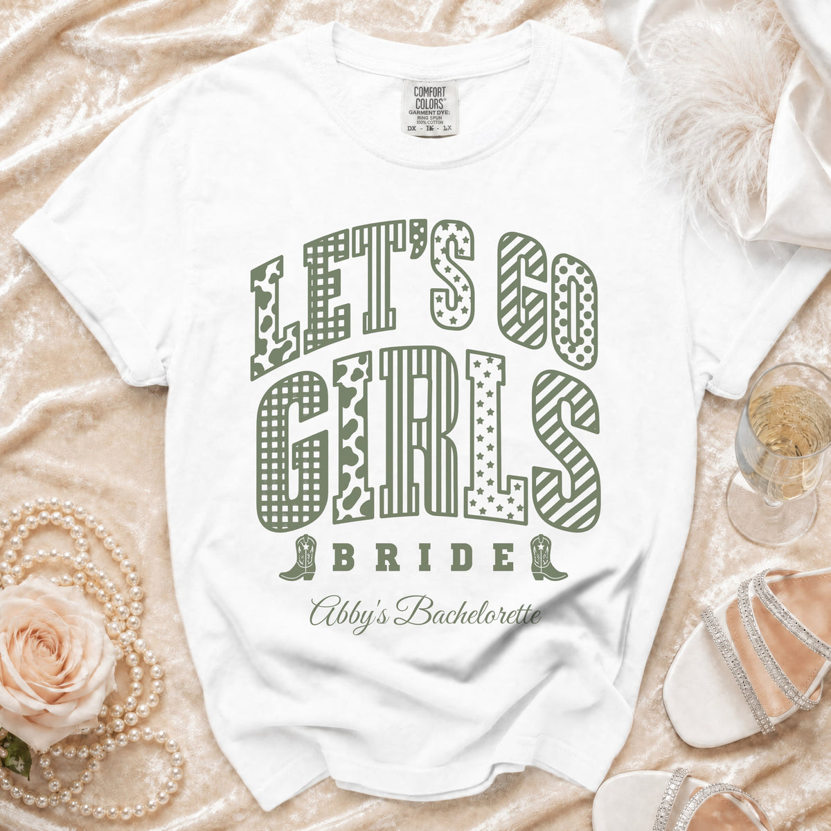 A white t-shirt features green text reading "LET'S GO GIRLS" and "Abby's Bachelorette" on a draped fabric background with accessories.