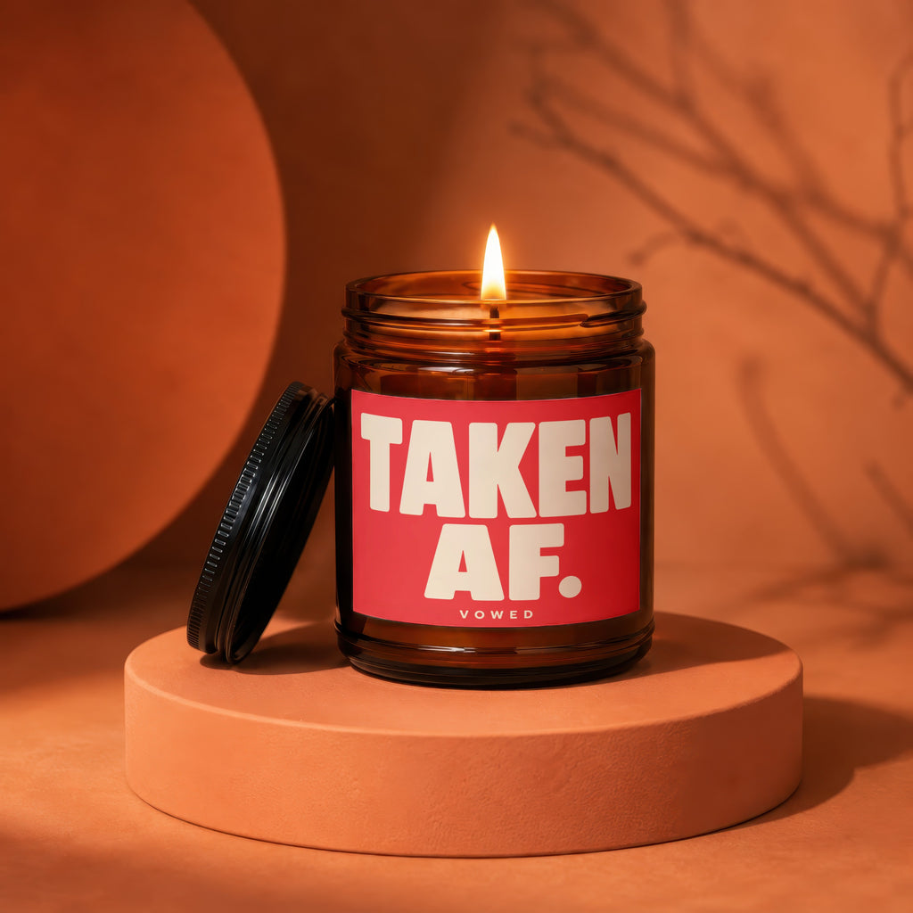 Brown glass candle with a lit wick and a red label reading "TAKEN AF. VOWED".