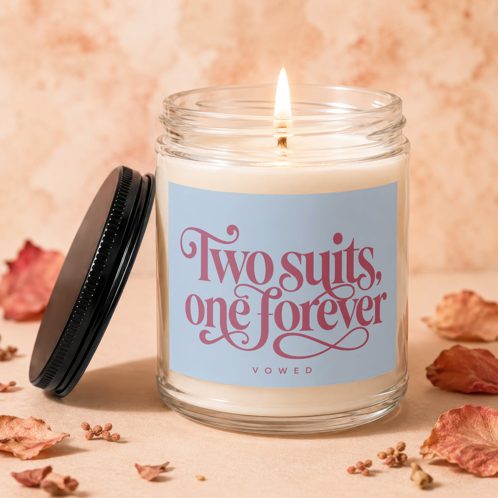 Glass jar candle with lit wick, reads "Two suits, one forever" and "VOWED".