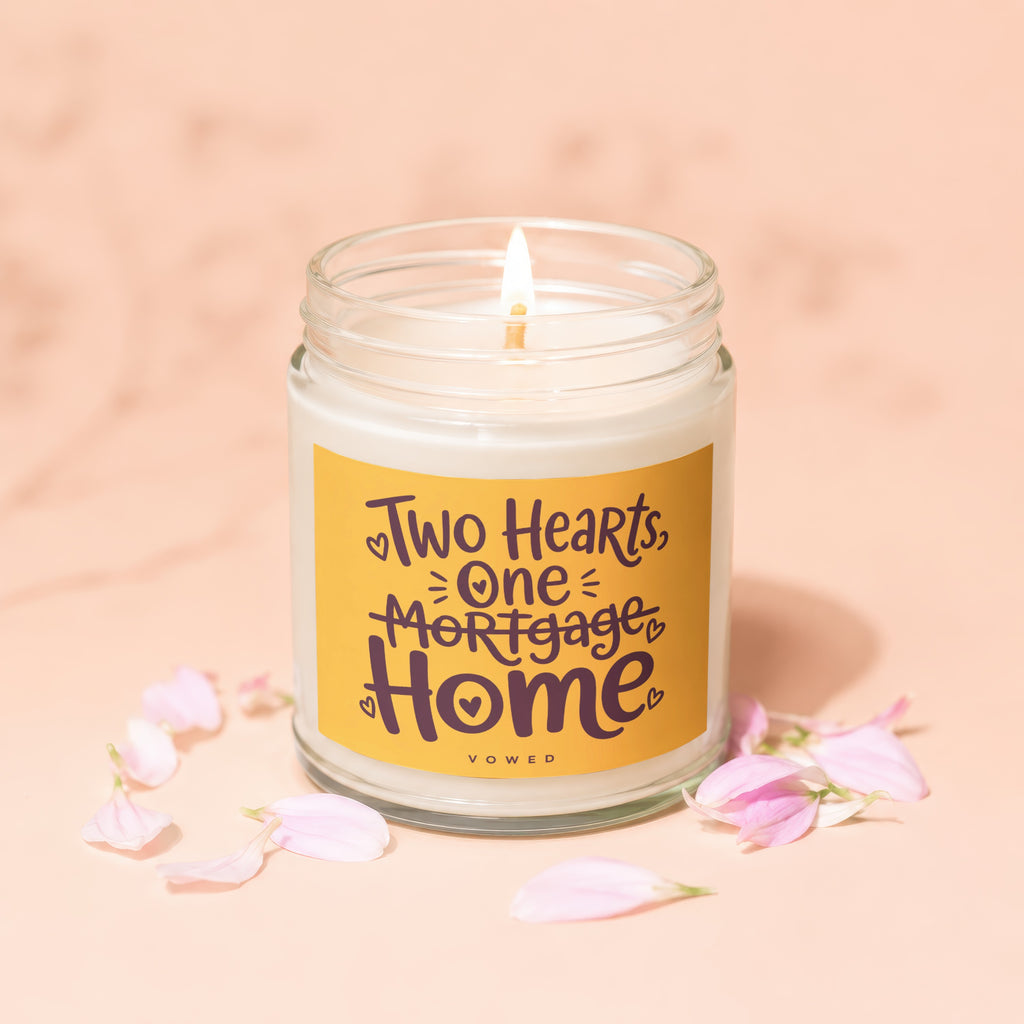 Glass jar candle with lit wick, featuring text "Two Hearts, One Mortgage, Home".